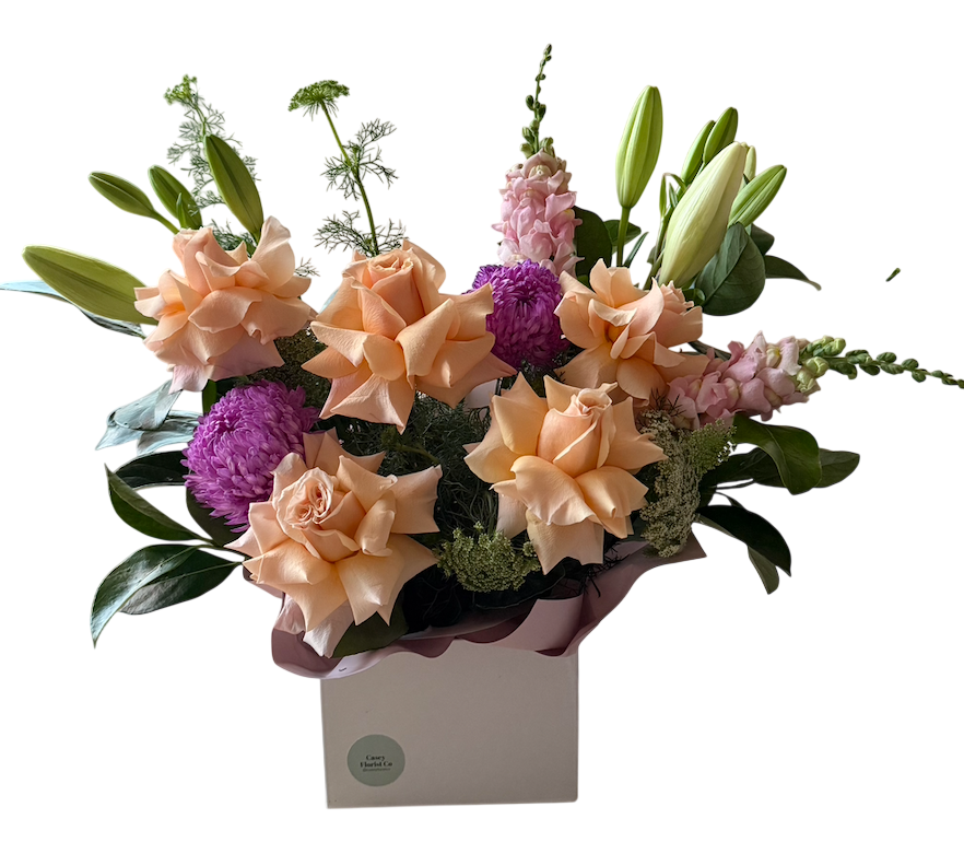 A floral arrangement featuring peach roses, purple chrysanthemums, pink stock, and green lily buds in a square white box with a dusty rose ribbon, set against a plain white background.